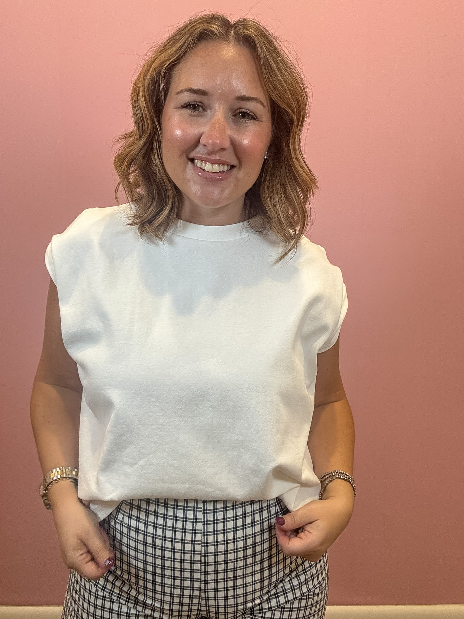A smiling woman with shoulder-length blonde hair wearing a white sleeveless crewneck t-shirt and a black-and-white gingham patterned skirt, standing against a solid pink wall.