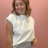 A woman with shoulder-length blonde hair smiling and looking down, wearing a white sleeveless crewneck top and a black and white gingham patterned skirt, standing against a solid pink wall.