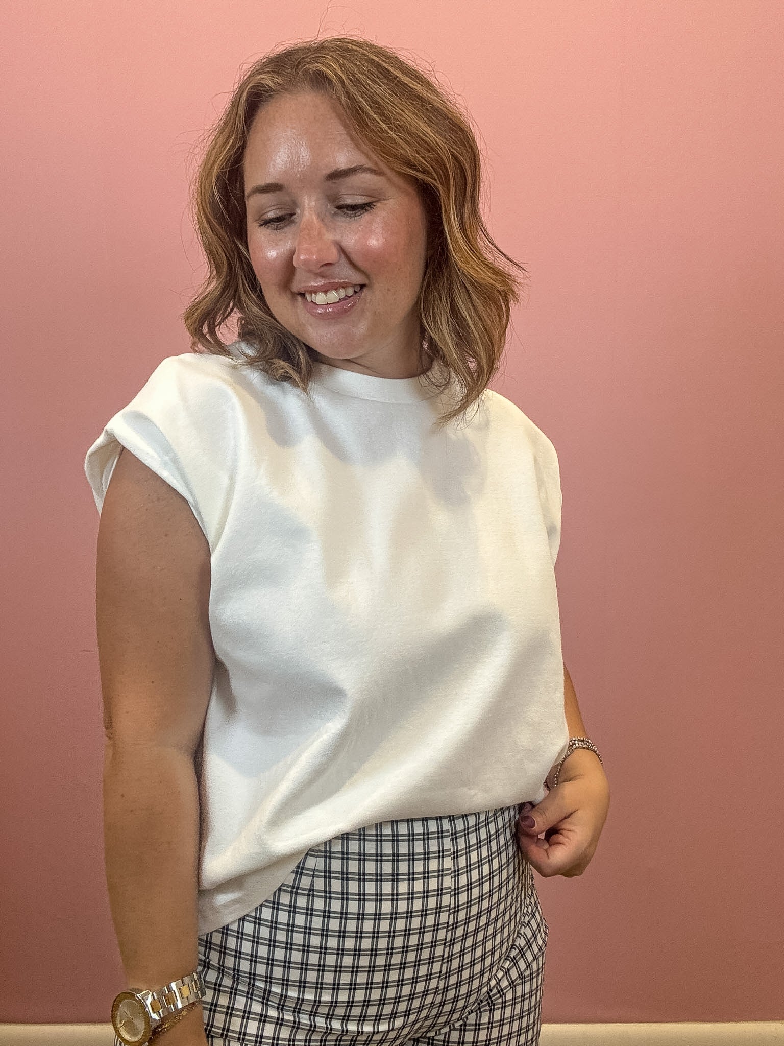 A woman with shoulder-length blonde hair smiling and looking down, wearing a white sleeveless crewneck top and a black and white gingham patterned skirt, standing against a solid pink wall.