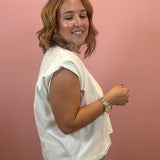 A woman with shoulder-length blonde hair is shown from the side, wearing a white sleeveless top and a black-and-white gingham skirt, standing against a solid pink wall.