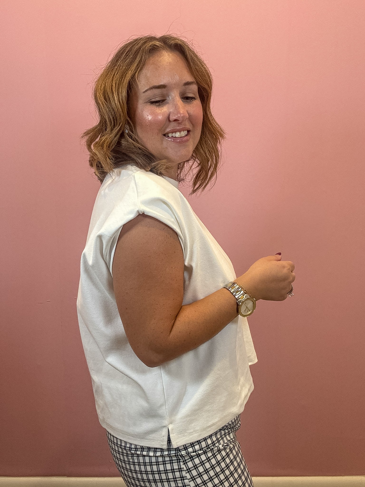 A woman with shoulder-length blonde hair is shown from the side, wearing a white sleeveless top and a black-and-white gingham skirt, standing against a solid pink wall.