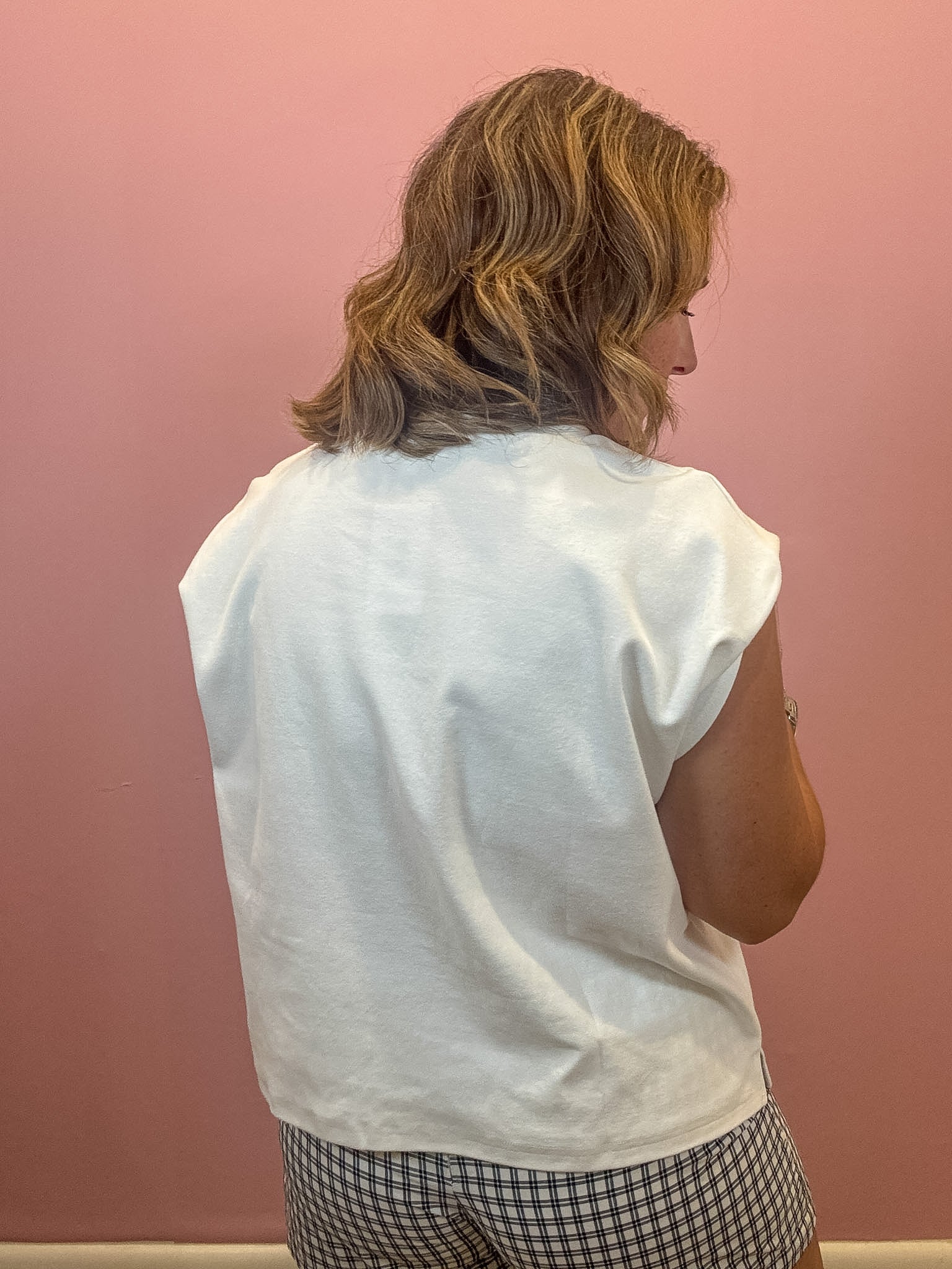 A woman viewed from behind wearing a white sleeveless top and a black-and-white gingham skirt, standing against a solid pink wall.