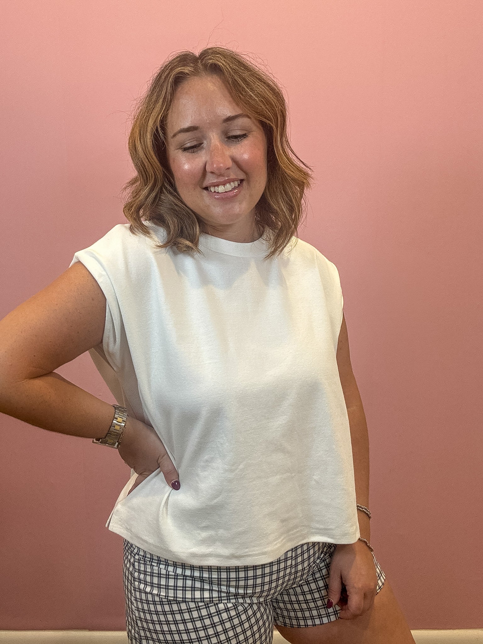 A woman wearing a white sleeveless crop top and black-and-white gingham shorts, standing against a solid pink wall.