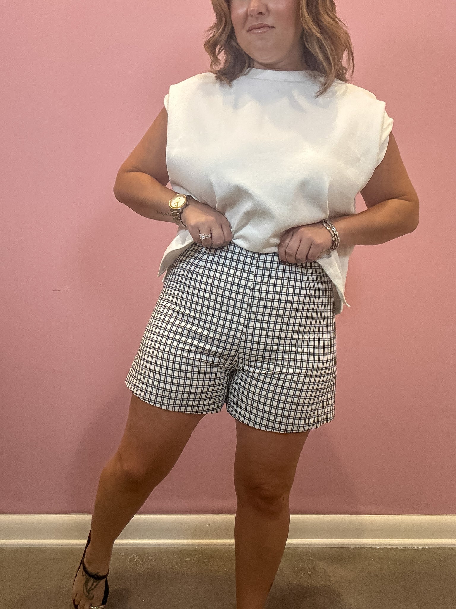 A woman modeling a white sleeveless top and gray-and-white gingham shorts against a solid pink wall.