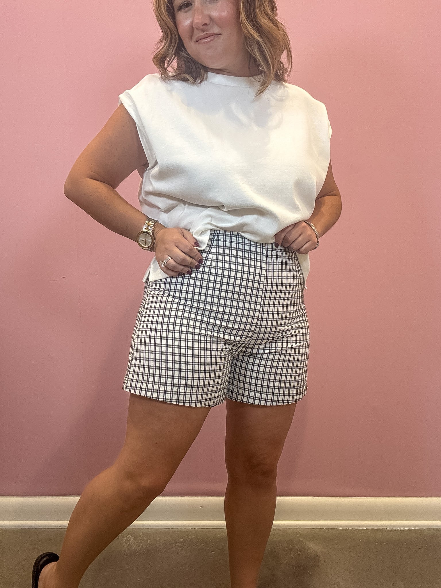 A woman modeling a white sleeveless top and gray and white gingham shorts against a solid pink wall.