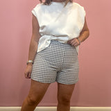 A woman modeling a white sleeveless t-shirt and gray and white gingham check shorts, standing against a solid pink wall.