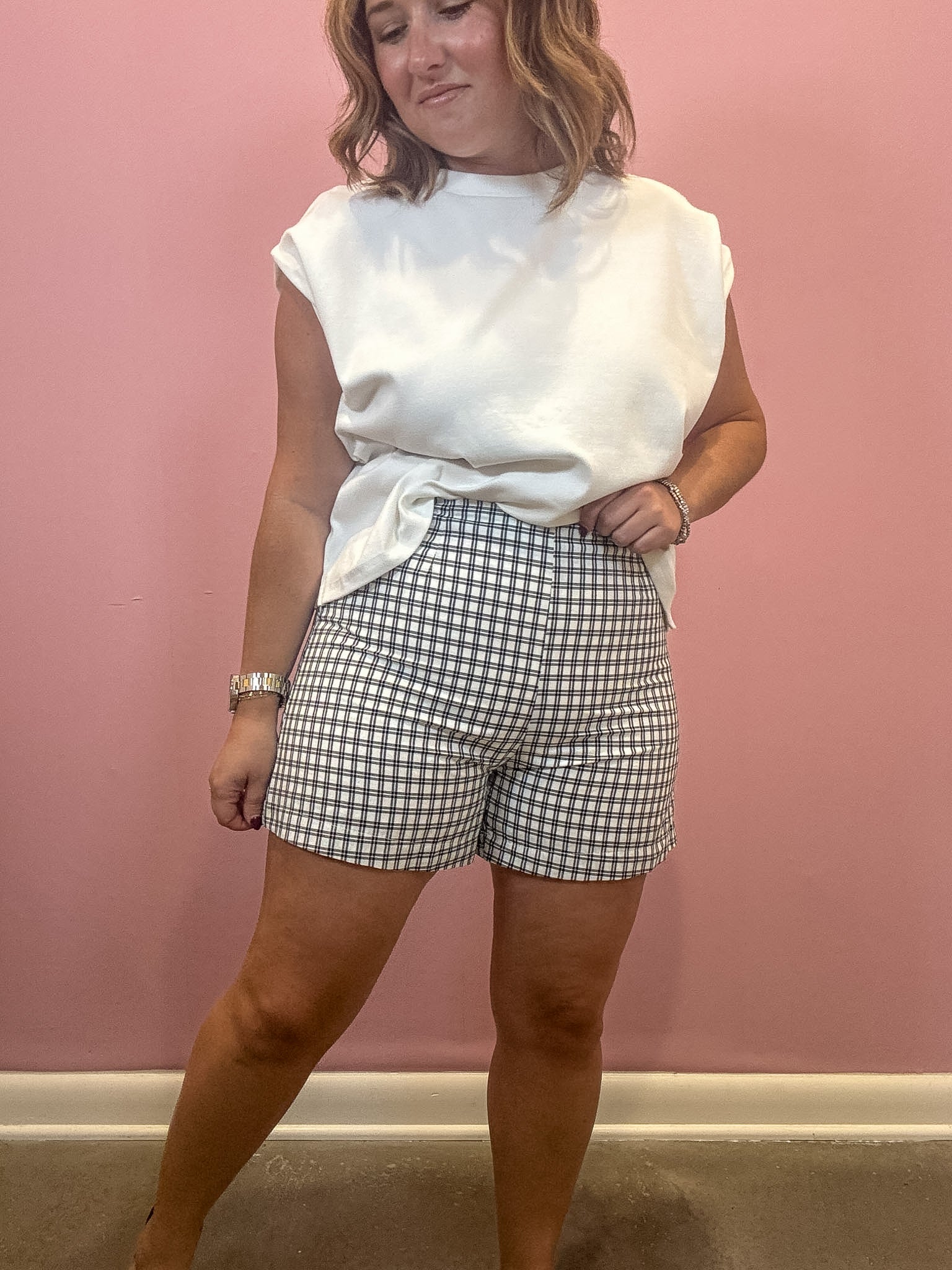 A woman modeling a white sleeveless t-shirt and gray and white gingham check shorts, standing against a solid pink wall.