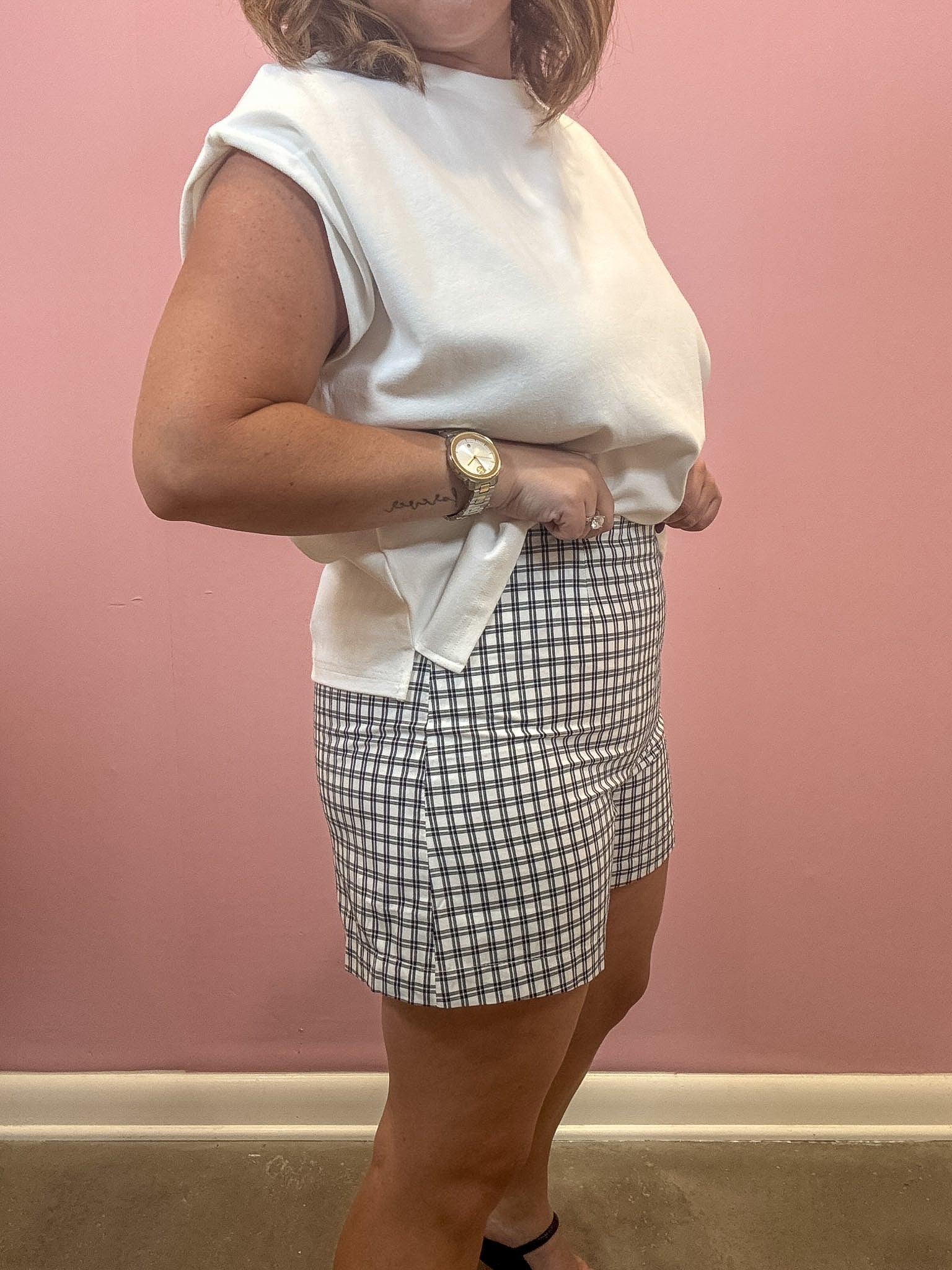 A woman wearing a white sleeveless top and a black and white gingham mini skirt, standing against a pink wall.
