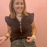 A woman smiling while wearing a dark brown ruffle-sleeve top with a button placket and patterned pants, standing against a solid pink wall.