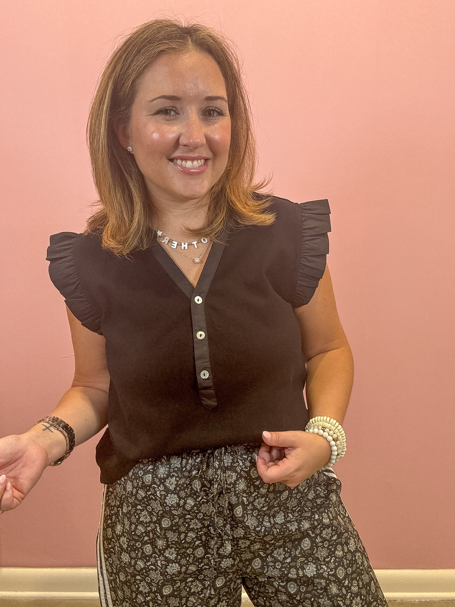 A woman smiling while wearing a dark brown ruffle-sleeve top with a button placket and patterned pants, standing against a solid pink wall.