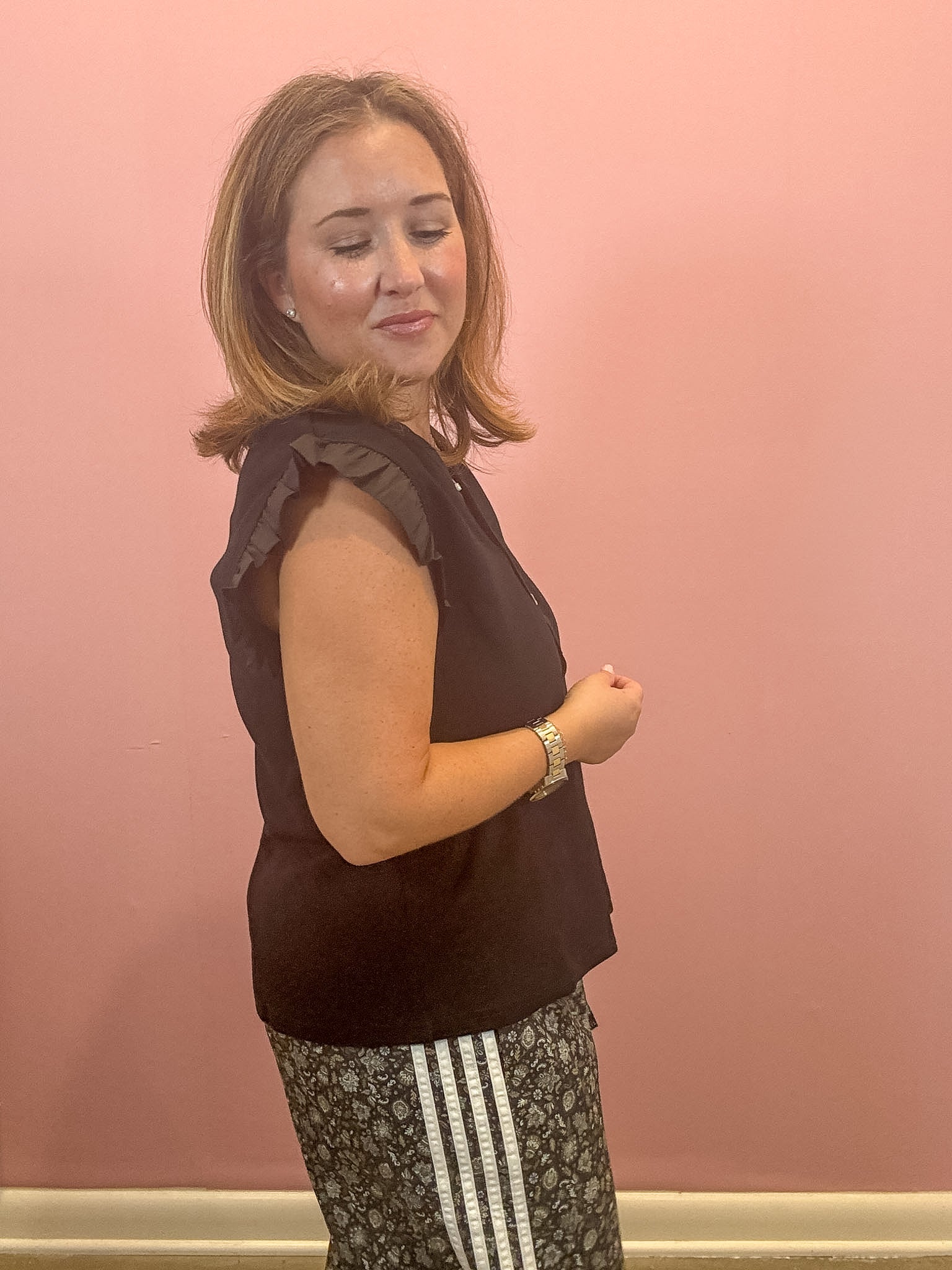 A woman wearing a black sleeveless top with ruffled shoulders and patterned pants with white stripes, standing against a pink wall.