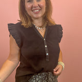 A smiling woman wearing a dark brown sleeveless blouse with ruffled shoulders and a button placket, paired with a patterned skirt, standing against a solid pink wall.