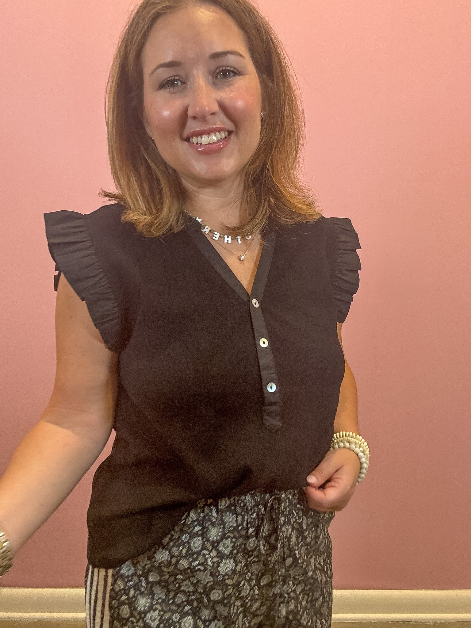 A smiling woman wearing a dark brown sleeveless blouse with ruffled shoulders and a button placket, paired with a patterned skirt, standing against a solid pink wall.