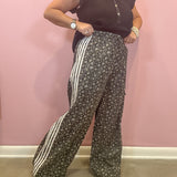 A woman modeling wide-leg pants with a dark gray and white floral pattern and three white side stripes, paired with a black ruffled sleeveless top, standing against a pink wall.