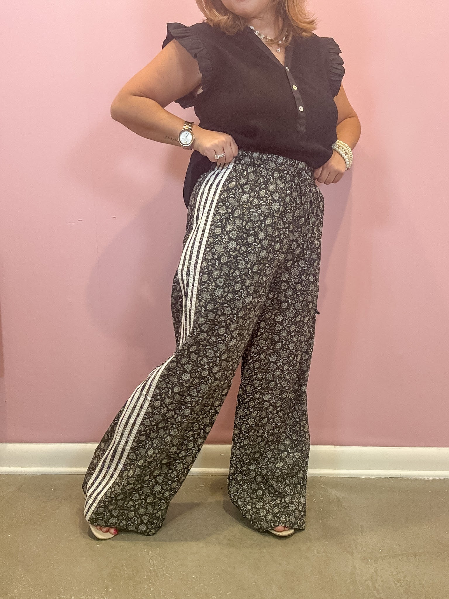 A woman modeling wide-leg pants with a dark gray and white floral pattern and three white side stripes, paired with a black ruffled sleeveless top, standing against a pink wall.