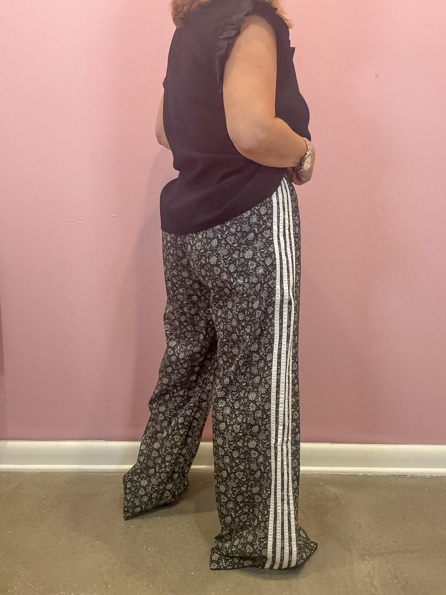 A person shown from the back wearing wide-leg patterned pants with three white side stripes, a dark sleeveless top, and a watch, standing against a pink wall.