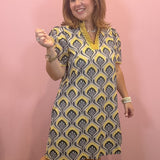 A woman modeling a short-sleeve V-neck dress with a yellow, gray, and white geometric pattern, standing against a solid pink wall.