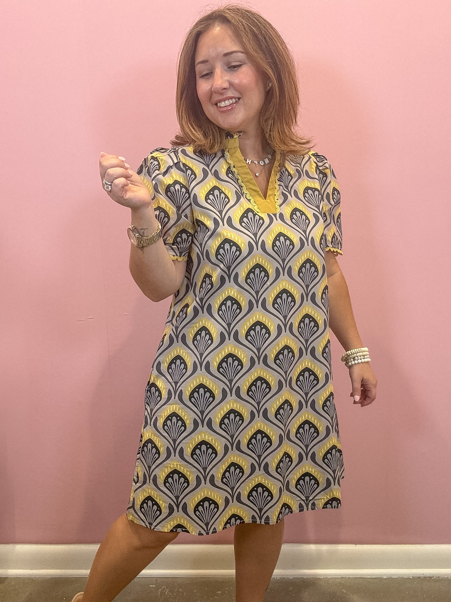 A woman modeling a short-sleeve V-neck dress with a yellow, gray, and white geometric pattern, standing against a solid pink wall.