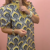A woman wearing a short-sleeve tunic top with a gray, yellow, and black geometric fan pattern, standing against a solid pink wall.
