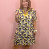 A woman modeling a short-sleeve V-neck dress with a yellow, gray, and white geometric pattern, standing against a solid pink wall.