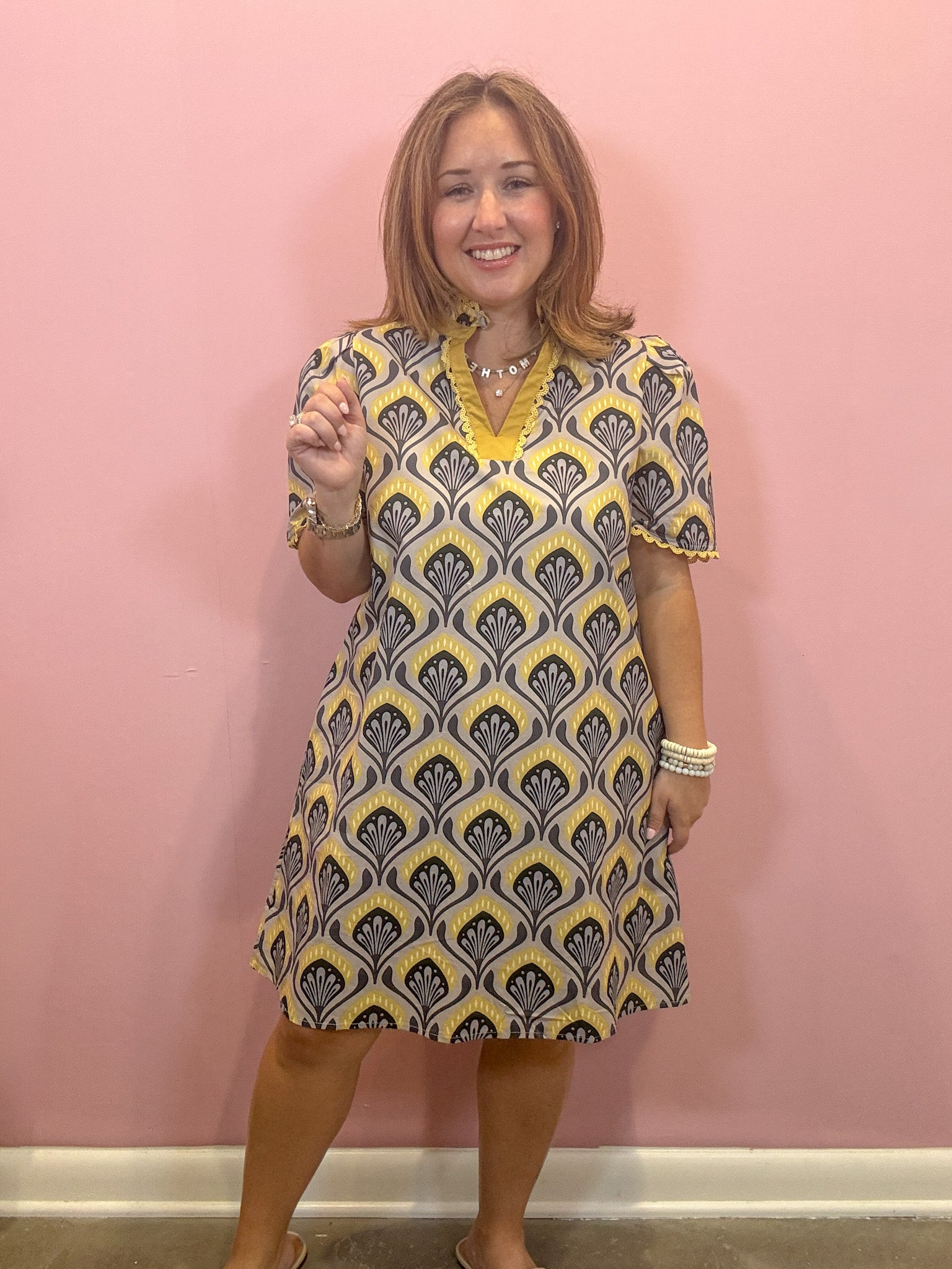 A woman modeling a short-sleeve V-neck dress with a yellow, gray, and white geometric pattern, standing against a solid pink wall.