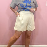 A woman modeling a short-sleeve striped t-shirt with a blue palm tree graphic and cream-colored scalloped shorts, standing against a pink wall.