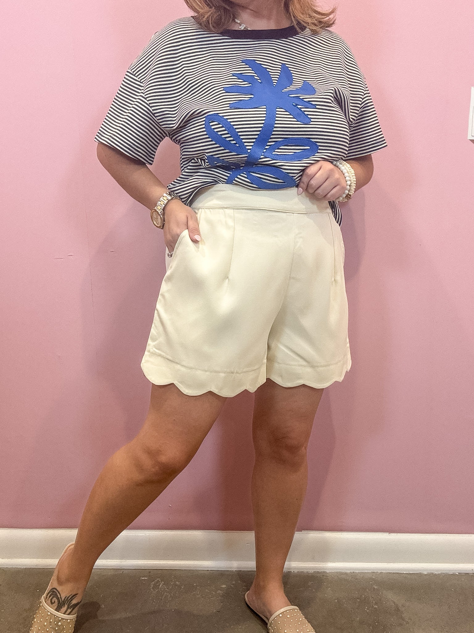 A woman modeling a short-sleeve striped t-shirt with a blue palm tree graphic and cream-colored scalloped shorts, standing against a pink wall.