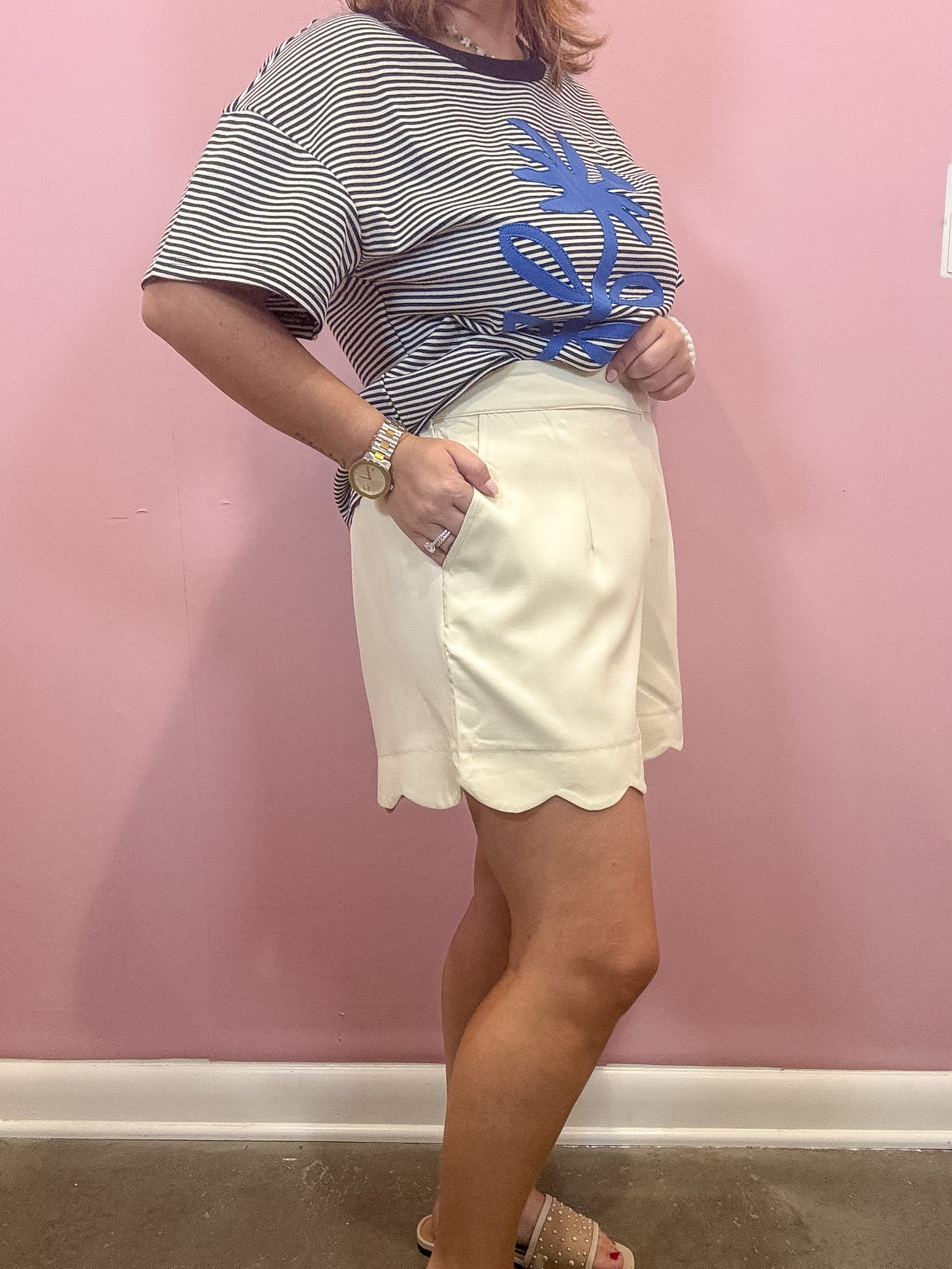A woman modeling a black and white striped t-shirt with a blue graphic and a cream-colored skirt, standing against a pink wall.