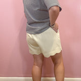 A person shown from the back wearing a gray and white striped short-sleeve top and cream-colored scalloped shorts, standing against a pink wall.