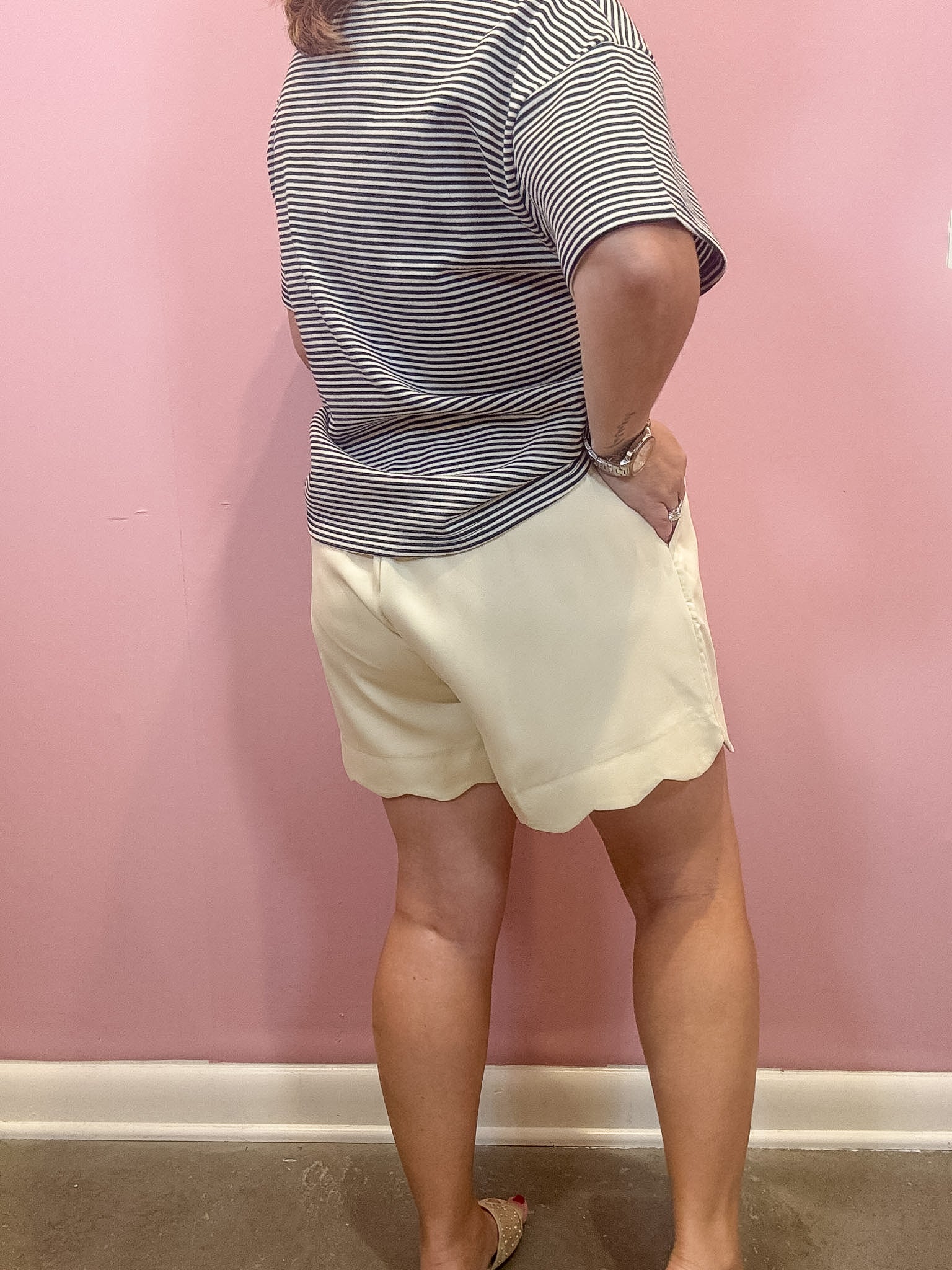 A person shown from the back wearing a gray and white striped short-sleeve top and cream-colored scalloped shorts, standing against a pink wall.