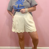 A woman modeling a short-sleeve striped t-shirt with a blue palm tree graphic and cream-colored scalloped shorts, standing against a pink wall.