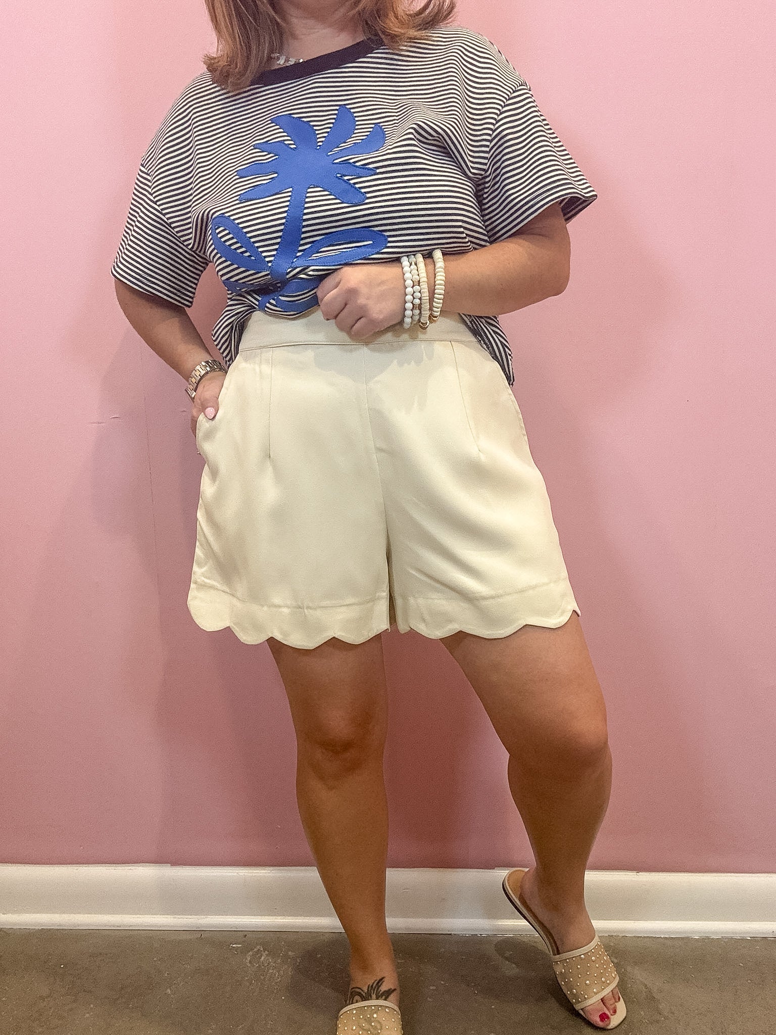 A woman modeling a short-sleeve striped t-shirt with a blue palm tree graphic and cream-colored scalloped shorts, standing against a pink wall.