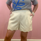 A person wearing cream-colored shorts with a scalloped hem and a navy and white striped t-shirt with a blue palm tree graphic, standing against a pink wall.