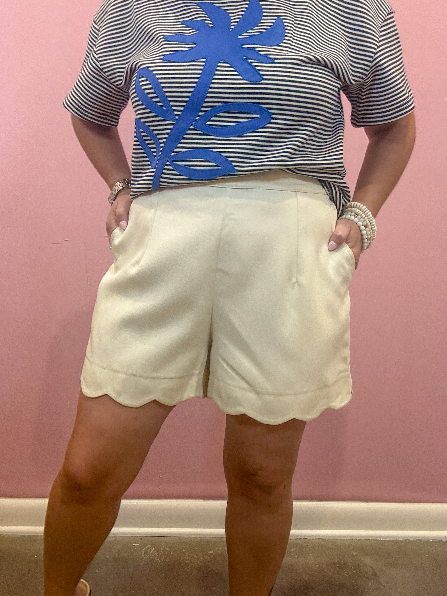 A person wearing cream-colored shorts with a scalloped hem and a navy and white striped t-shirt with a blue palm tree graphic, standing against a pink wall.