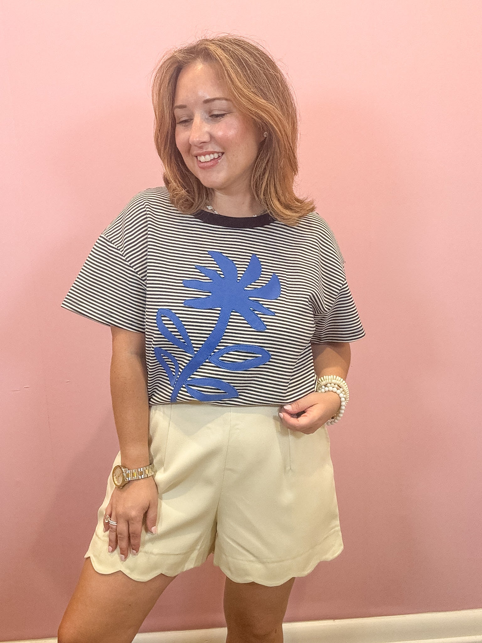 A woman modeling a short-sleeve striped t-shirt with a blue palm tree graphic, paired with cream scalloped shorts, standing against a solid pink wall.