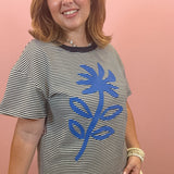 A woman wearing a black and white horizontally striped t-shirt with a large blue palm tree graphic on the front, paired with beige shorts, standing against a solid pink wall.