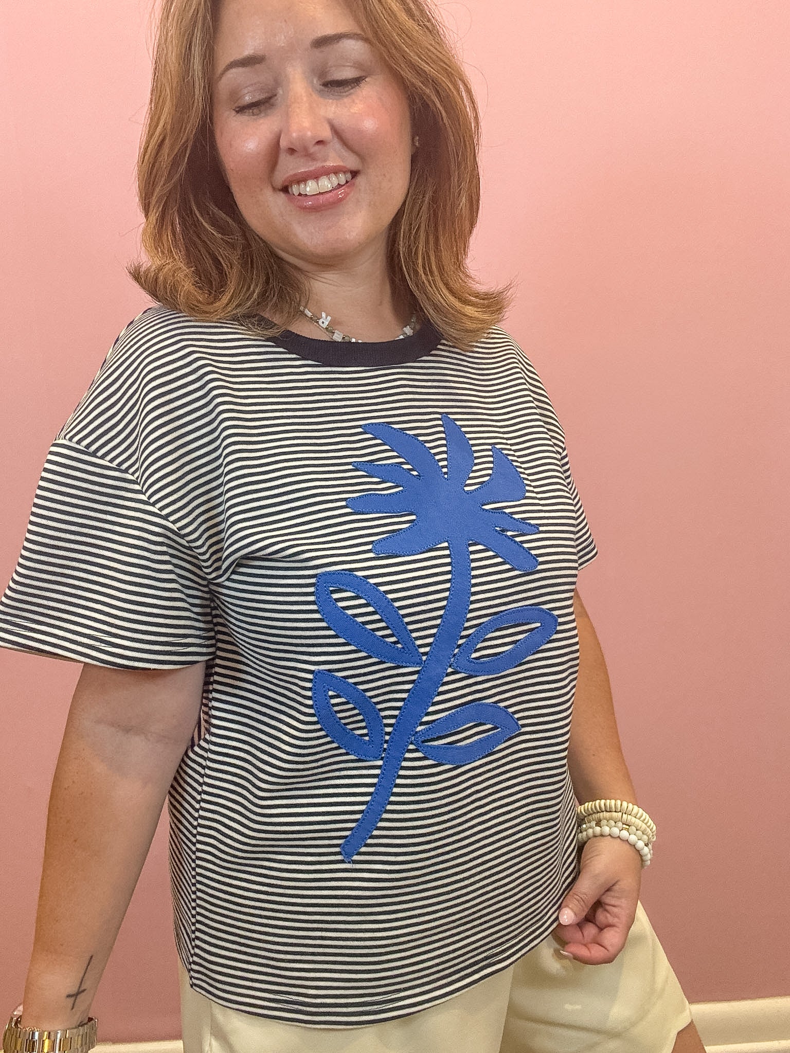 A woman wearing a black and white horizontally striped t-shirt with a large blue palm tree graphic on the front, paired with beige shorts, standing against a solid pink wall.