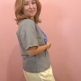 A woman with shoulder-length blonde hair stands against a pink wall, wearing a black and white horizontally striped short-sleeve top with a blue graphic print on the front, paired with cream-colored shorts.