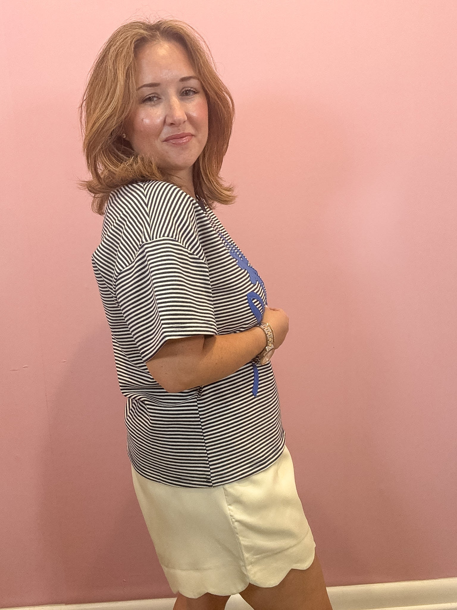 A woman with shoulder-length blonde hair stands against a pink wall, wearing a black and white horizontally striped short-sleeve top with a blue graphic print on the front, paired with cream-colored shorts.