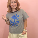 A woman modeling a black and white striped t-shirt with a blue abstract graphic on the front, paired with cream scalloped shorts, standing against a solid pink wall.