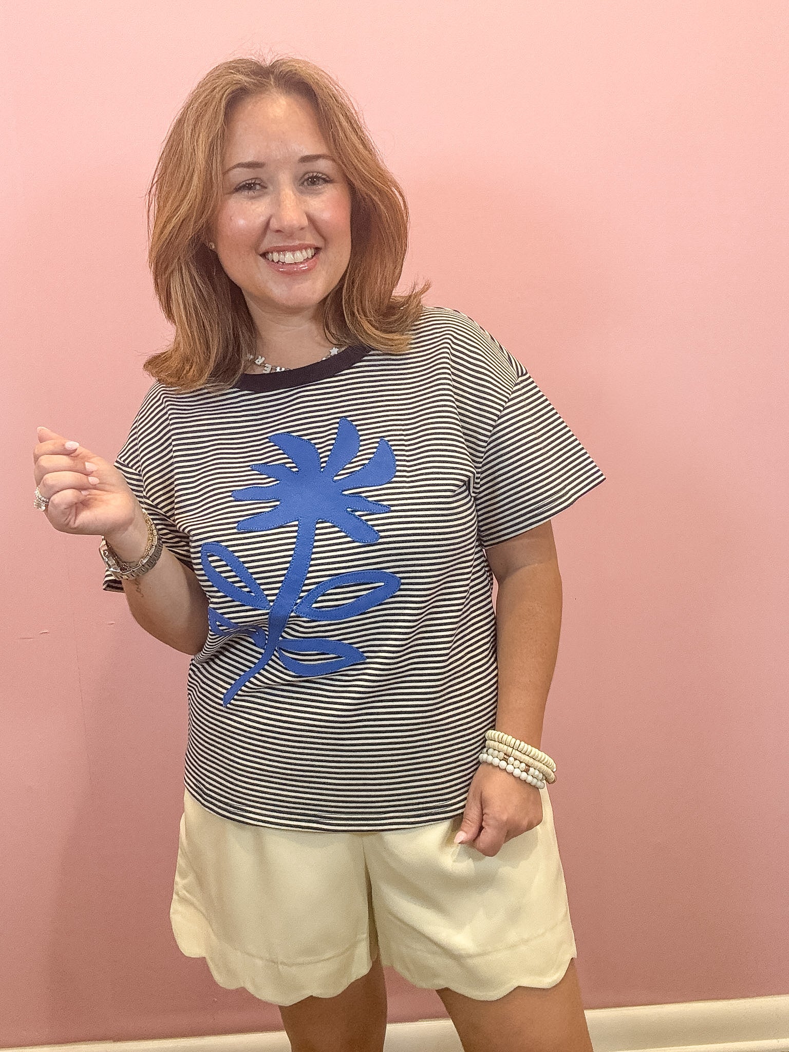 A woman modeling a black and white striped t-shirt with a blue abstract graphic on the front, paired with cream scalloped shorts, standing against a solid pink wall.