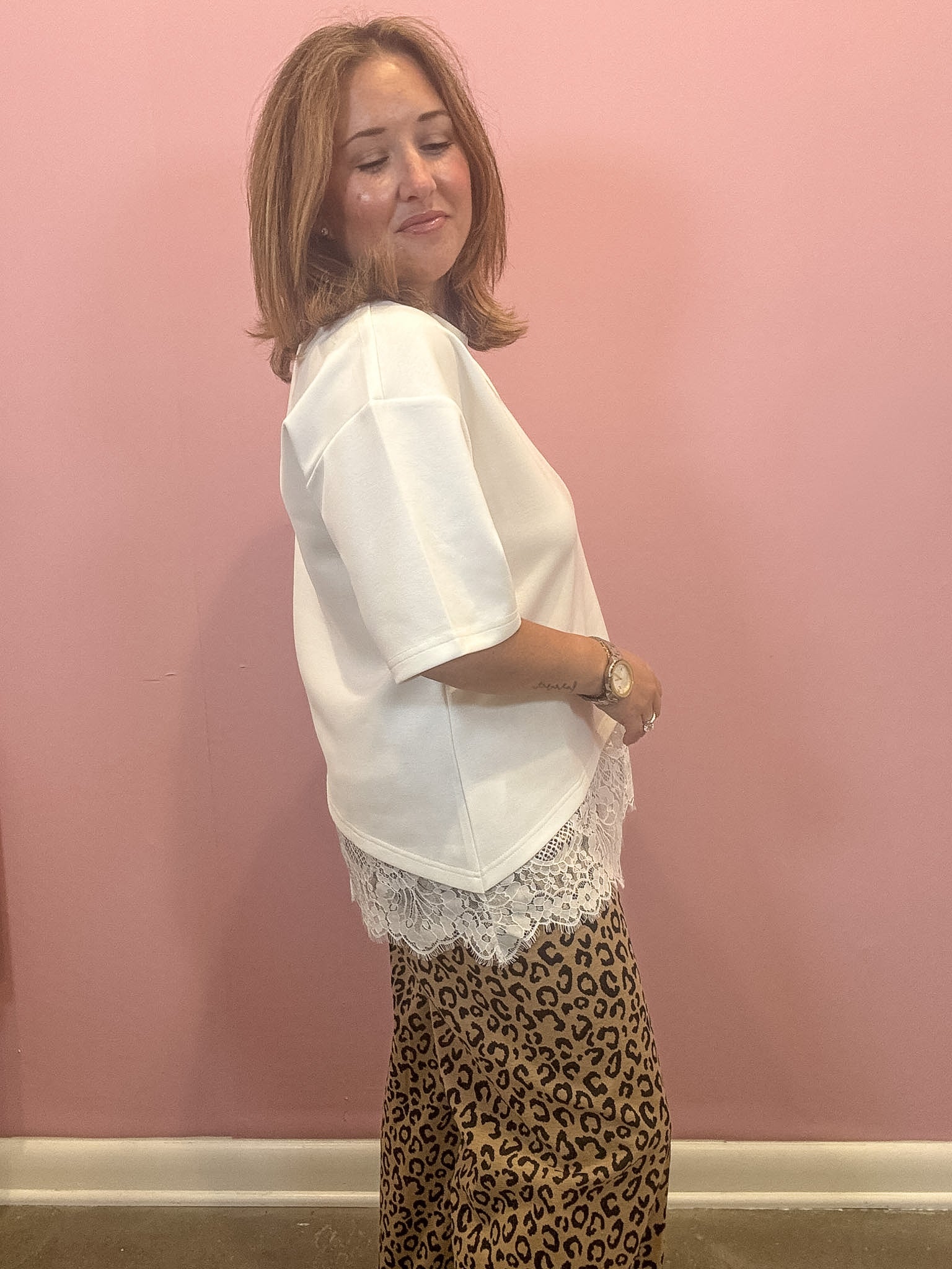 A woman wearing a white short-sleeve blouse with a lace hem and leopard-print pants, standing against a pink wall.