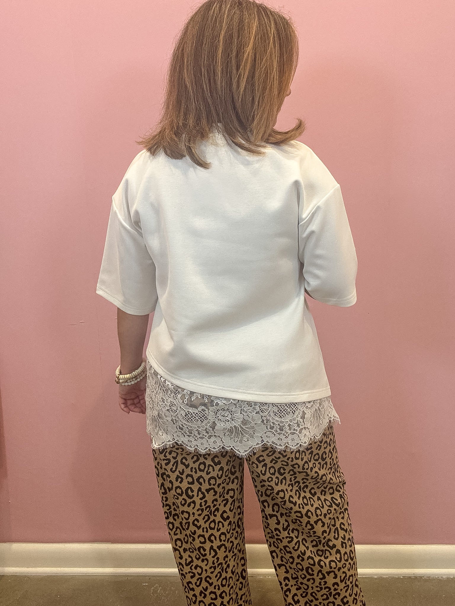 A person shown from the back wearing a white short-sleeve top with a lace hem, paired with leopard-print pants, standing against a pink wall.