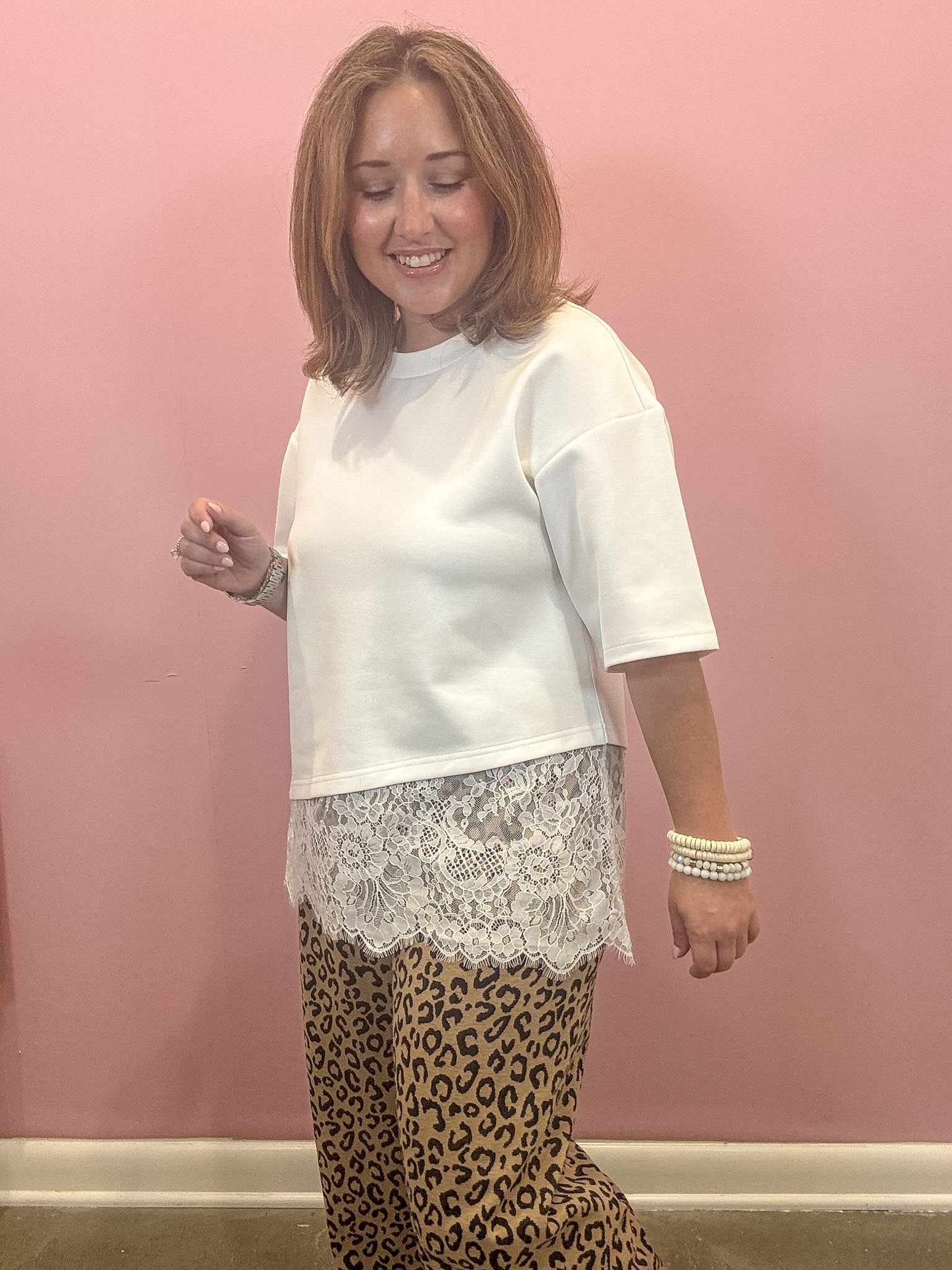 A woman modeling a white short-sleeve crewneck top with a white lace hem, paired with leopard print pants, standing against a solid pink wall.