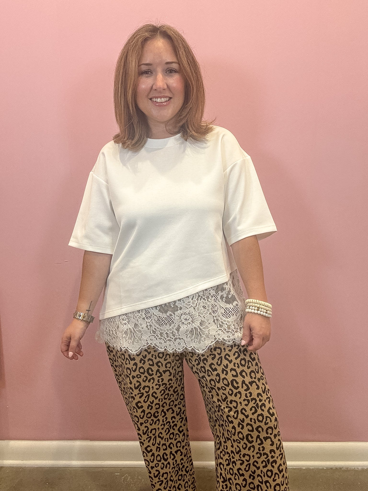 A woman modeling a white short-sleeve t-shirt with a sheer white lace hem overlay, paired with leopard print pants, standing against a solid pink wall.