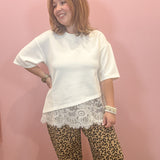 A woman modeling a white short-sleeve top with a white lace hem, paired with leopard print pants, standing against a solid pink wall.