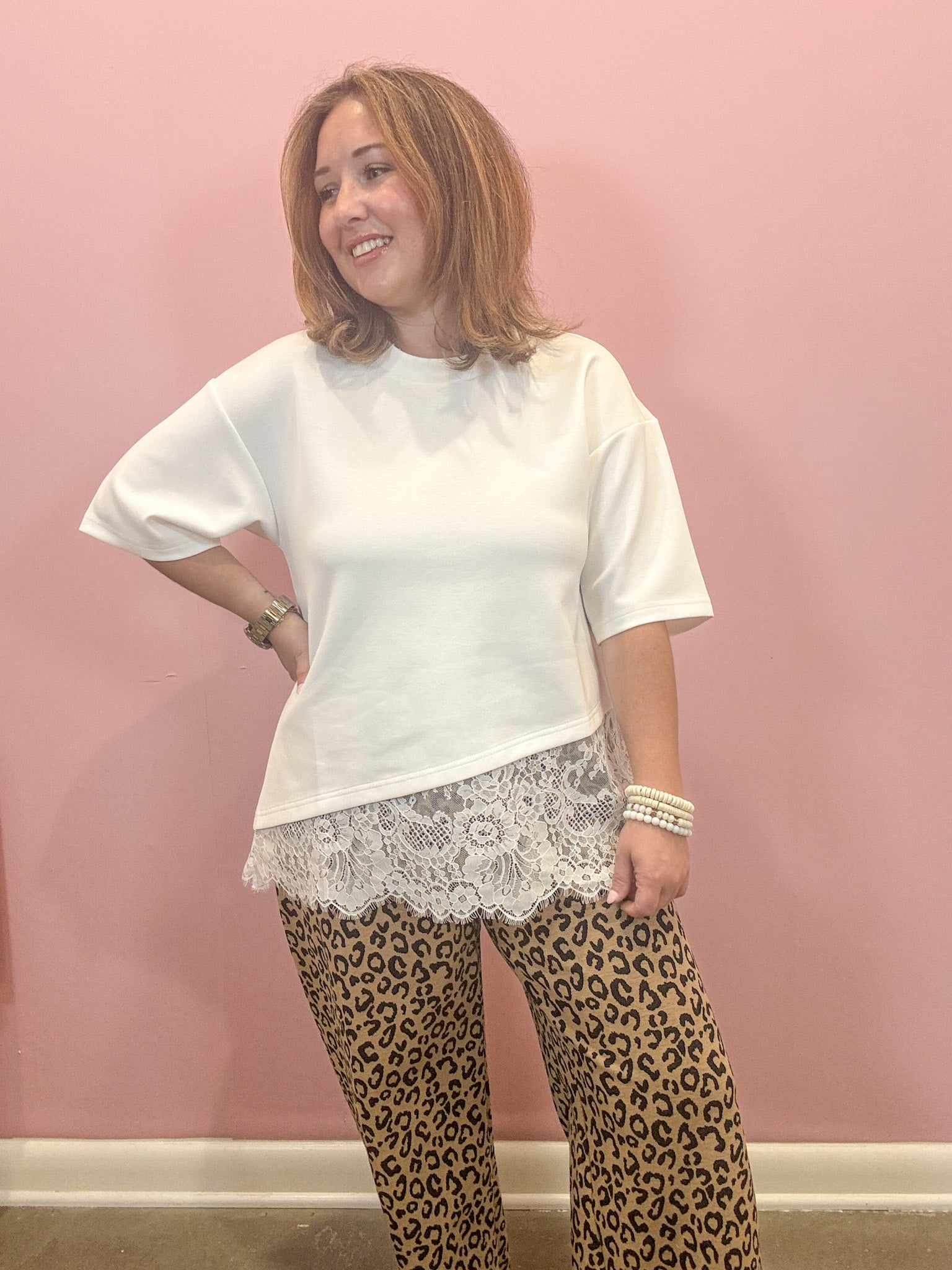 A woman modeling a white short-sleeve top with a white lace hem, paired with leopard print pants, standing against a solid pink wall.