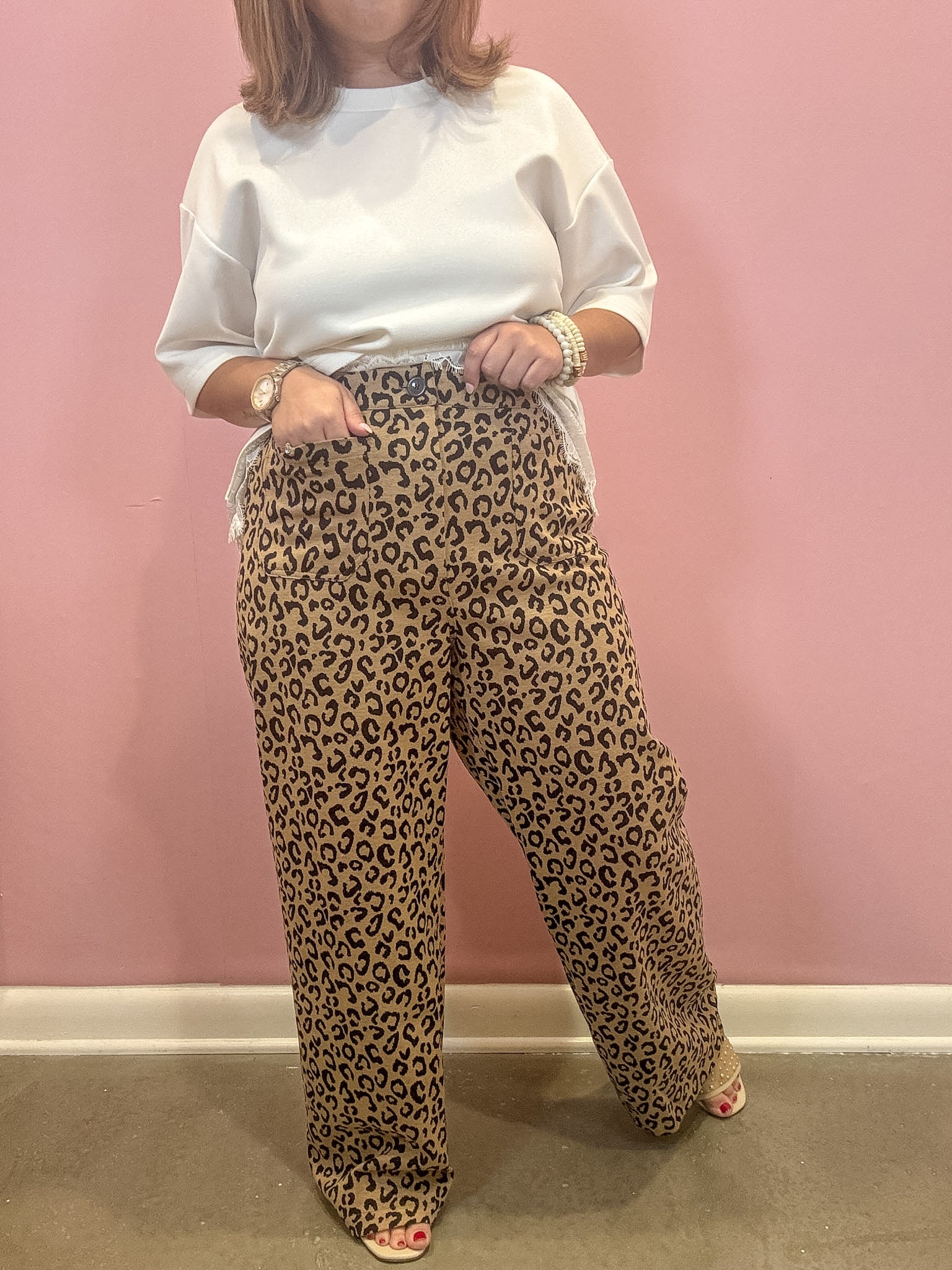 A person wearing leopard-print wide-leg pants and a white top stands against a pink wall, with the pants as the likely product focus.