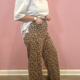 A person wearing loose leopard-print wide-leg pants and a white short-sleeve top, standing against a pink wall.