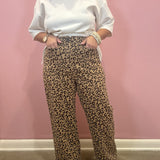 A person wearing loose leopard-print pants and a white sweatshirt stands against a pink wall, with hands in the pockets of the pants.